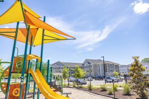 The playground area of The Sullivan, a new grey three story apartment building, is in the foreground on a sunny day with clear blue skies in the background.