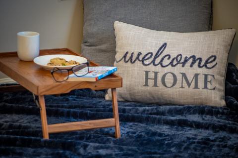 A food tray is set to the left, and two pillows are to the right. All are sitting on a blanket. The front pillow is emblazoned with the message "welcome home."